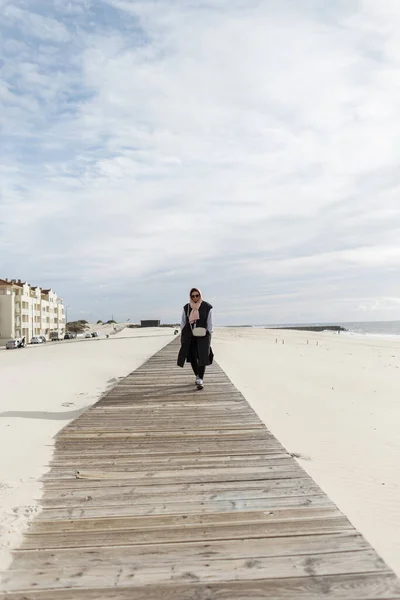 Fashionable beautiful woman with a scarf on her head wearing a vest, sweatshirt, and sneakers walks on a wooden walkway on the beach in the ocean