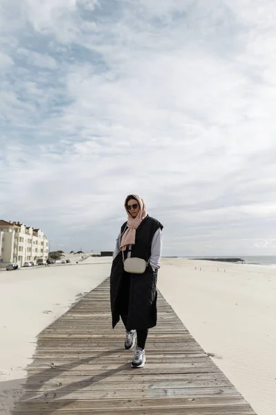 Stylish beautiful woman with a scarf on her head and sunglasses wearing a spring fashionable black vest, sweatshirt, handbag and sneakers walks along a wooden walkway on the beach by the ocean.