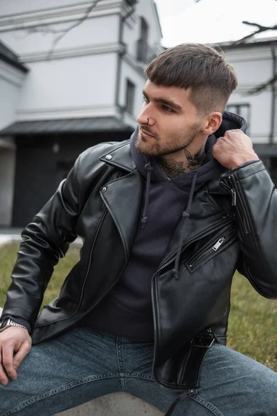 Fashionable handsome man with hair and a beard with a tattoo on his neck in fashionable casual outfit with a hoodie and a jacket sits on the street
