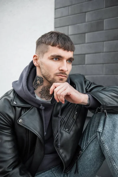 Fashionable handsome model man hipster with hairstyle and beard with a tattoo on his neck in a black leather jacket and hoodie sitting on the street