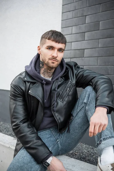 Fashion handsome hipster guy with hair and tattoos on his neck wearing trendy casual clothes with a black leather jacket, stylish hoodie, jeans and sneakers sitting outside near a black brick wall