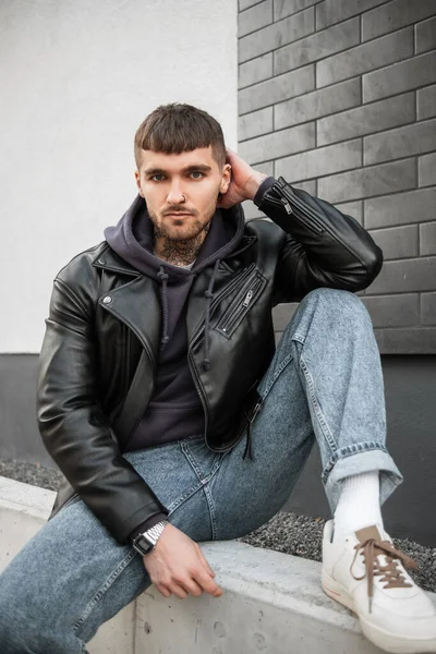 Stylish handsome fashionable man model hipster with hair and beard with a tattoo on his neck in a black leather jacket, hoodie, jeans and white sneakers sits on the street near a black brick wall