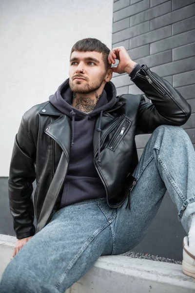 Fashion hipster handsome man with tattoos on neck in casual clothes with leather jacket, hoodie and jeans sits and poses near a white and brick brick wall on the street