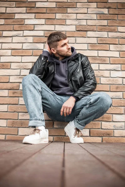 Fashionable handsome young man hipster model in a stylish outfit with leather jacket, hoodie, jeans and white sneakers sitting near a brick wall