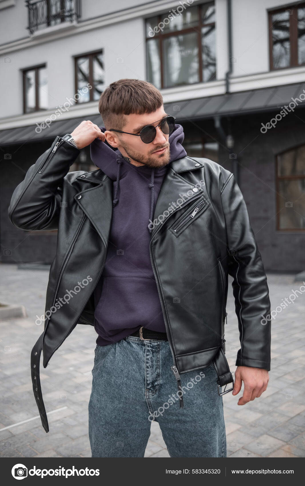 El hombre modelo elegante y guapo de moda con ropa de moda con chaqueta de  cuero y gafas de sol lleva una capucha y camina por la ciudad