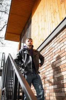 Tuğla duvarın yanındaki merdivenlerde duran kapüşonlu ve kotlu şık deri ceketli şık, genç bir erkek model.