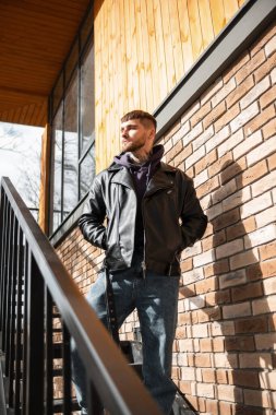 Tuğla duvarın yanındaki merdivenlerde duran kapüşonlu ve kotlu şık deri ceketli şık, genç bir erkek model.