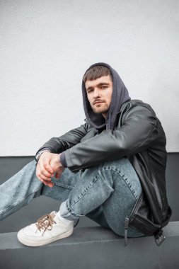 Saçlı ve sakallı yakışıklı hippi ceketli, kapüşonlu, kot pantolonlu ve spor ayakkabılı adam sokakta beyaz duvarın yanında oturuyor.