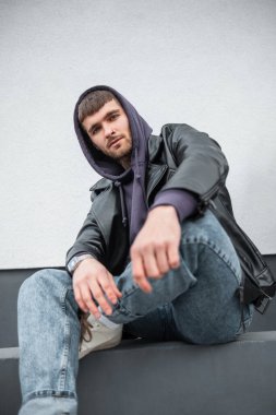 Havalı moda modasına uygun yakışıklı erkek modeli, rock moda deri ceketli, kapüşonlu ve kot pantolonlu sokakta beyaz bir duvarın yanında oturuyor.