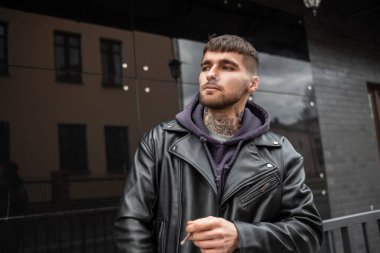 Deri ceketli ve kapüşonlu yakışıklı genç hippi bir sigara alır ve caddedeki siyah bir binanın yanında yürür.