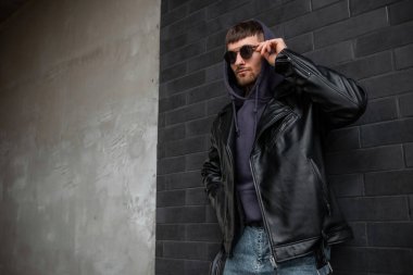 Cool handsome man model in fashionable clothes with leather rock jacket and hoodie is wearing stylish vintage sunglasses and standing near a black brick wall on the street