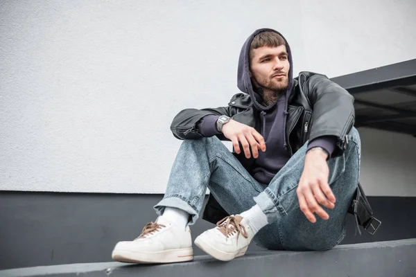 Cool street style man hipster in fashion casual wear with trendy rock leather jacket, hoodie, jeans and white sneakers sitting near a white wall on the street