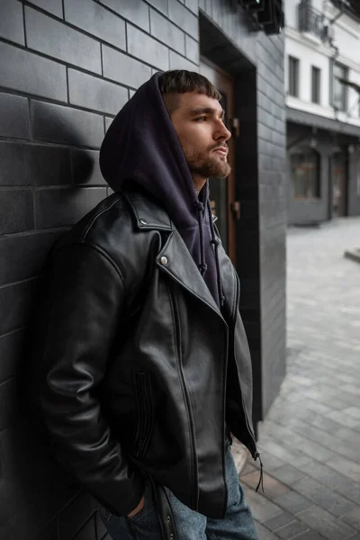 Stylish street handsome man in a fashionable black rock leather jacket with a purple hoodie stands near a black brick building on the street