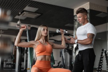 Dövmeli yakışıklı vücut geliştirme eğitmeni güzel sarışın bir kadını eğitiyor ve spor salonunda nasıl egzersiz yapılacağını gösteriyor. Dambıllarla spor yapan kız.