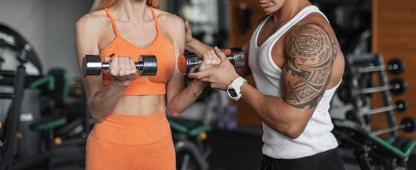 Women\'s hands holding metal dumbbells and pumping her biceps with a fitness trainer. Guy helps a girl work out at the gym