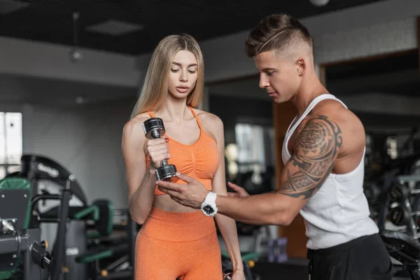 Sexy pretty blonde woman with a sportswear is workout and trains with a personal trainer at the gym. Fitness girl and bodybuilder guy work out with dumbbells