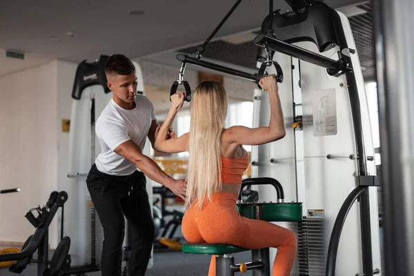 Spor salonunda birlikte çalışan güzel bir atletik çift. Seksi vücutlu güzel sarışın atletik kadın egzersiz makinesine oturur ve sırt kaslarını spor hocasıyla doldurur.