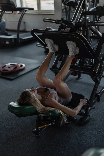 穿着时髦运动服 穿着运动鞋 体形性感的漂亮的运动健美姑娘躺在模拟器上 在体育馆里锻炼腿肌肉 — 图库照片