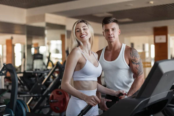 Gülümseme egzersizi yapan ve spor salonunda birlikte antrenman yapan güzel ve sağlıklı bir çift. Güzel sarışın fitness kadını. Moda spor kıyafetleriyle gülümseyen. Koşu bandında spor hocasıyla koşan.