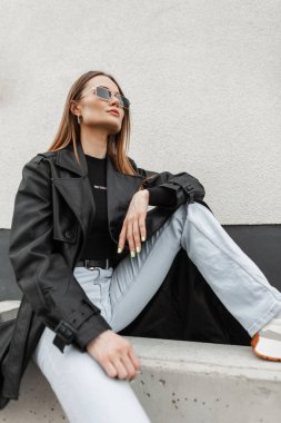 Şık güneş gözlüklü, modaya uygun giysileri olan, kotlu ve tişörtlü şık deri ceketli güzel bir kadın mankeni sokakta oturuyor.