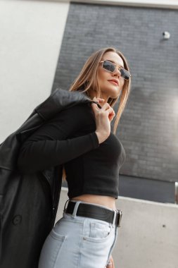 Havalı, altın rengi gözlüklü, siyah tişörtlü, kot pantolonlu ve paltolu şık bir kız sokakta yürüyor.
