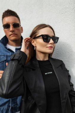 Moda gözlüklü, modaya uygun siyah bir ceket ve tişört giyen şık, genç ve güzel bir kadın model ve sokakta şık kot giyimli genç bir adam. Modaya uygun bir çift.