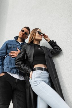 Şık, genç ve şık şehir kıyafetleri içinde, deri ceketli ve kot pantolonlu güzel bir çift duvarın yanında duruyor. Havalı moda güneş gözlüğü takan şık, güzel hippi kadın.