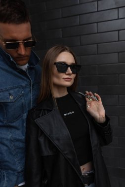 Moda gözlüklü, siyah modaya uygun kıyafetli, siyah tuğlalı bir duvarın yanında havalı bir hipsterle duran güzel bir kız.