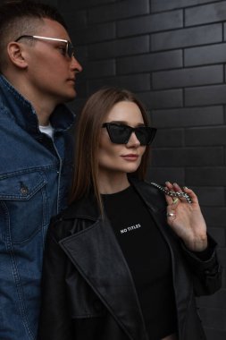 Moda güzeli çift kızı ve şık güneş gözlüklü şık yakışıklı adam şık şehir kıyafetleri içinde deri ceketli ve çantalı kot pantolon şehirdeki siyah tuğla duvarın yanında duruyor.