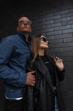 Güneş gözlüklü şık elbiseli havalı, şık, havalı hipster çift sokakta siyah tuğlalı bir duvarın yanında duruyor. Güzel bir kız ve modaya uygun giyinmiş yakışıklı bir erkek.