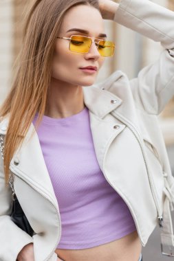 Beyaz deri ceketli, sokakta mor tişörtlü, şık güneş gözlüklü güzel bir kızın moda sokak portresi.