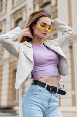 Mor tişörtlü ve yüksek bel kotlu sarı gözlüklü şık genç hippi kız şehirde moda olan beyaz deri ceketli.