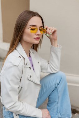 Moda derisi ceketli, şık, klasik kot pantolonlu güzel hippi kadın duvarın yanında oturuyor.