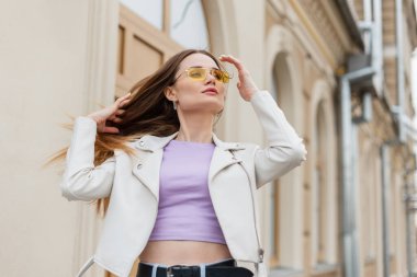 Şık, genç bir kız mankeni. Havalı, günlük kıyafetler içinde. Deri ceketli, mor tişörtlü, şehirde gezen ve saçını düzelten.
