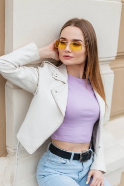 Moda modası geçmiş hippi güzel genç kadın modeller, şık günlük kıyafetlerle, deri ceketli, mor tişörtlü ve kot pantolonlu caddedeki klasik bir binanın yanında oturuyor.