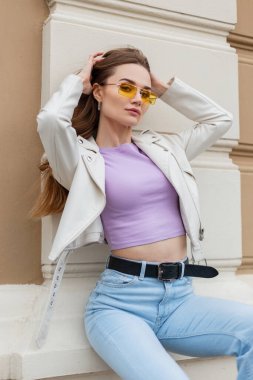 Beautiful sexy fashion model woman in fashionable bright outfit with a white leather jacket and vintage blue jeans with yellow sunglasses sits and poses near the wall by the building