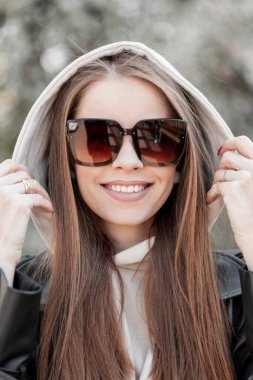 Women's fresh portrait of a beautiful stylish young happy woman model with a smile wearing fashionable sunglasses in casual outfit with a leather jacket and hoodie enjoying and walking in spring park