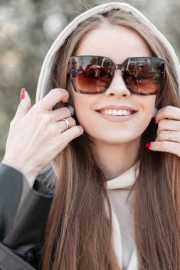 Fresh female portrait of a beautiful stylish young happy woman with a smile and cool vintage fashion sunglasses in a fashionable black leather jacket wearing a hood and walking in the city