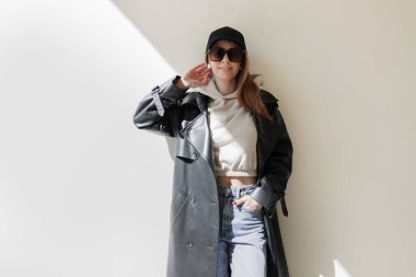Şapkalı, modaya uygun güneş gözlüklü, şık deri ceketli ve kot pantolonlu güzel model güneş ışığı olan bir duvarın yanında duruyor. Kadın şehir kıyafetleri.