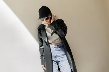 Cool fashion street beautiful woman hipster with trendy sunglasses and black cap in casual urban outfit with coat, hoodie and jeans stands near a wall