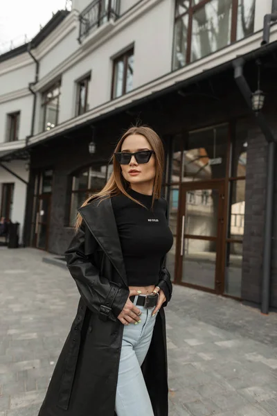 Beautiful trendy hipster girl with vintage sunglasses in trendy black clothes with black stylish mockup T-shirt with fashion black leather coat with jeans walking in the city