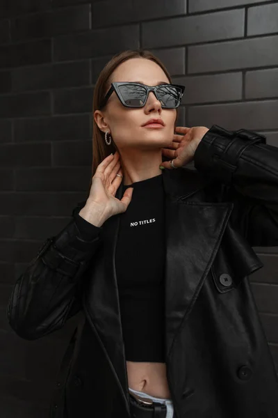 Fashion urban female portrait of beautiful girl with trendy sunglasses in black leather coat with black t-shirt stands and poses near a black brick background. Female style outfit , beauty and fashion