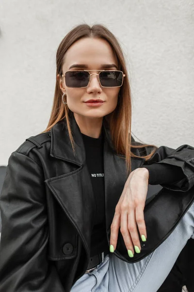 stock image Women's street fashionable portrait of a young beauty girl with trendy gold eyeglasses in fashion black outfit with a stylish leather coat and T-shirt sitting on the street