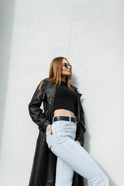 Glamour fashion street woman with vintage sunglasses in stylish black leather long coat with t-shirt and blue jeans stands near a white wall on the street