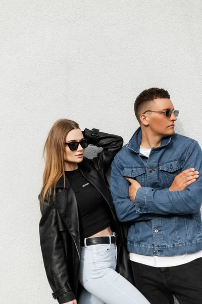 Fashionable beautiful young stylish couple woman and handsome man with vintage sunglasses in fashion clothes with jeans jacket and leather black coat standing near the wall. Trendy urban casual outfit