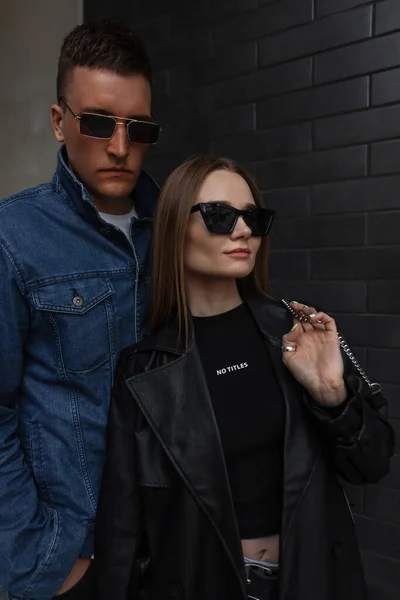 Fashionable street young couple beautiful girl and handsome man in fashionable clothes with sunglasses and purse are walking near the black brick wall in the city