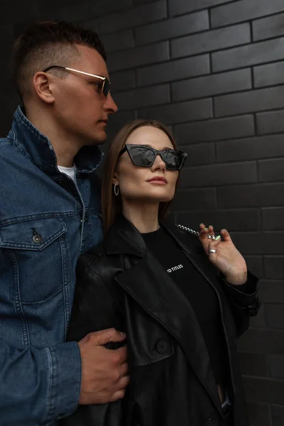 Fashionable trendy beautiful couple woman and man in fashion outfit with trendy sunglasses in black coat and jeans jacket stands and poses near a black brick wall on the street