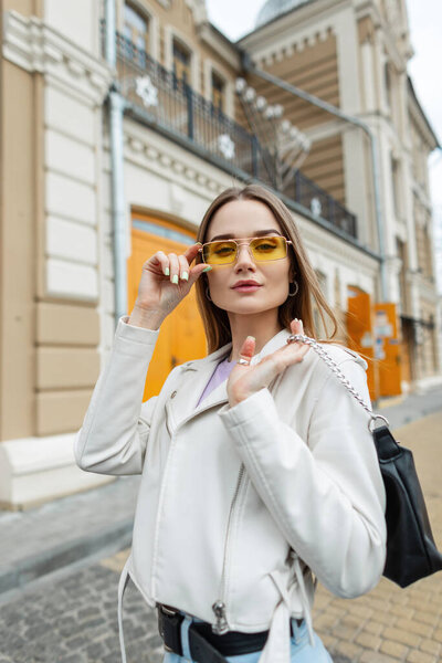 Fashionable beautiful woman model in fashion clothes with leather jacket and purse is wearing stylish vintage yellow glasses and walking in the city