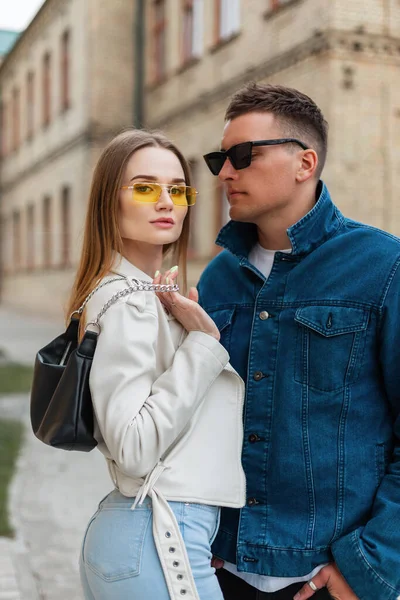 Fashion street beauty couple with sunglasses in trendy cool denim  outfit with leather jacket and jeans with bag stands in the city. Fashionable woman and hipster man