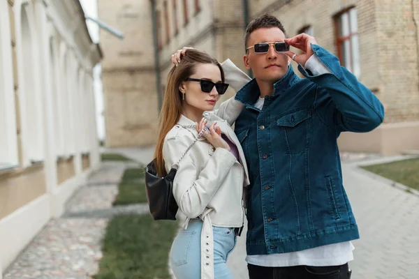 Stylish beautiful girl with a fashionable man in fashionable denim casual clothes with jacket, purse and sunglasses in the city. Fashion beauty couple models
